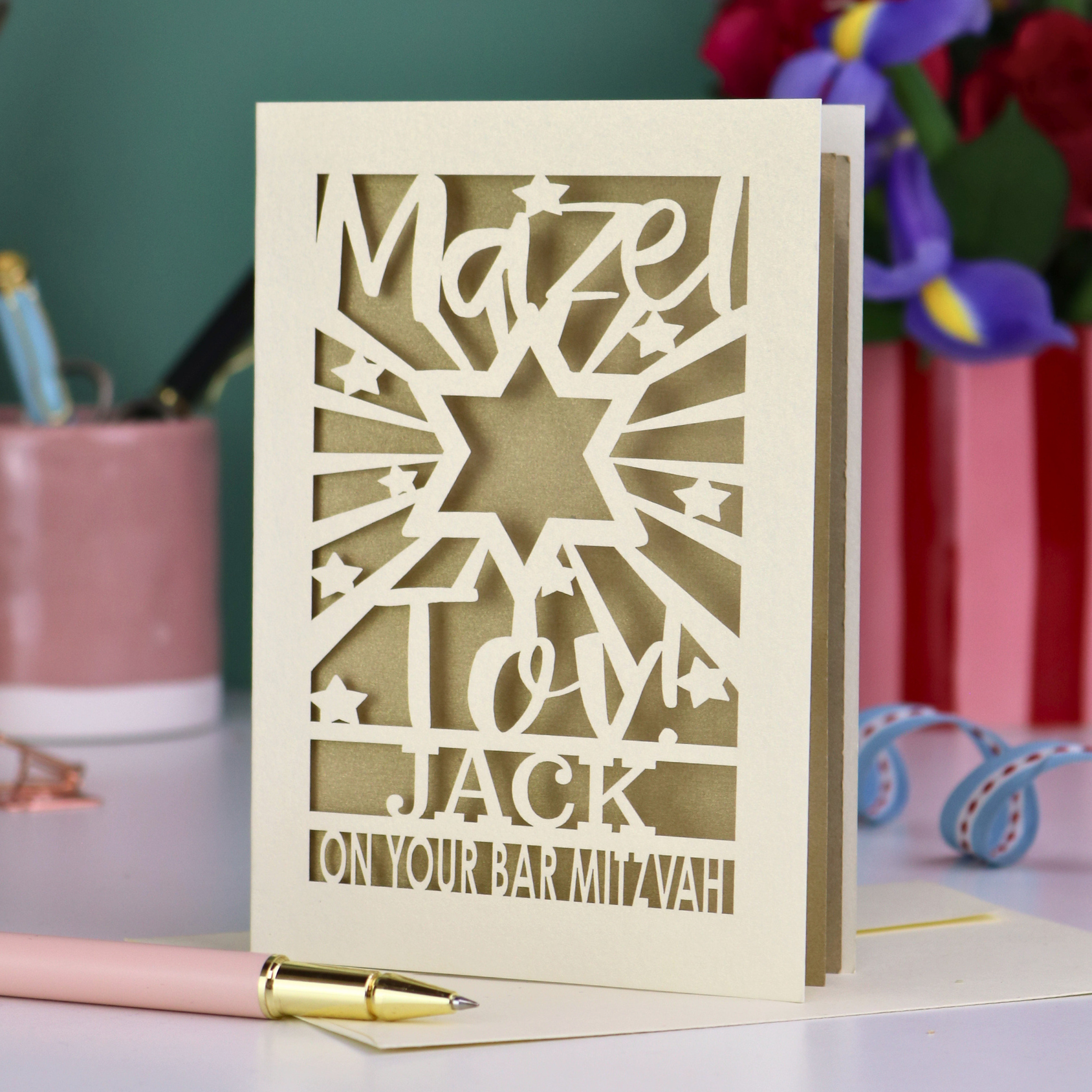 Decorative Laser cut Bar Mitzvah card with cut-out text, 'Mazel Tov Jack on your Bar mitzvah' on a table with stationery items.