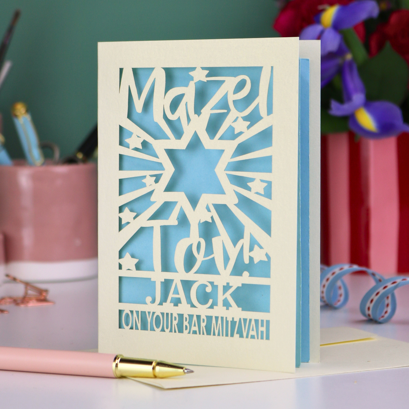 Decorative Laser cut Bar Mitzvah card with cut-out text, 'Mazel Tov Jack on your Bar mitzvah' on a table with stationery items.
