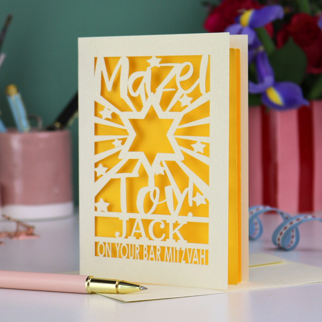 Decorative Laser cut Bar Mitzvah card with cut-out text, 'Mazel Tov Jack on your Bar mitzvah' on a table with stationery items.