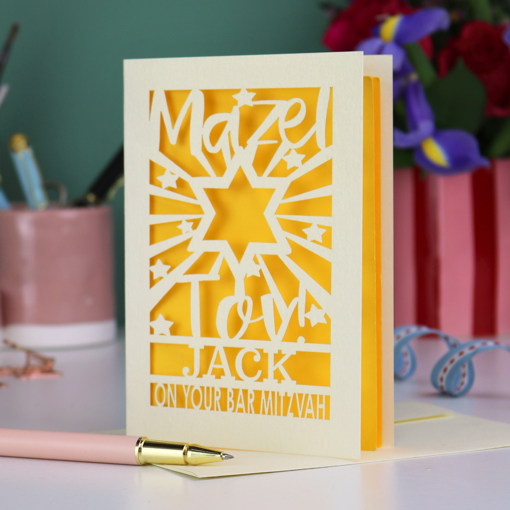 Decorative Laser cut Bar Mitzvah card with cut-out text, 'Mazel Tov Jack on your Bar mitzvah' on a table with stationery items.