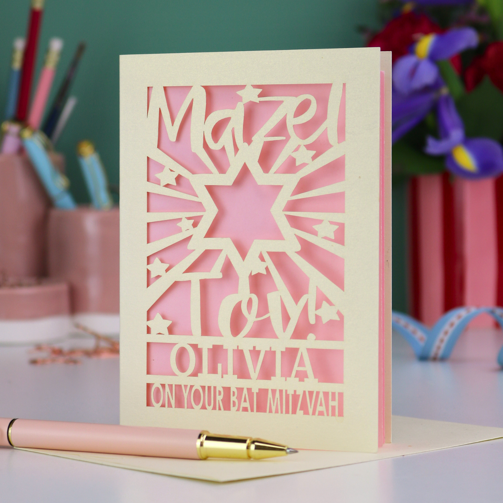 Personalized Bat Mitzvah card with decorative cut-out  star design and text 'Mazel Tov Olivia on your Bat Mitzvah' on a desk with stationery items.