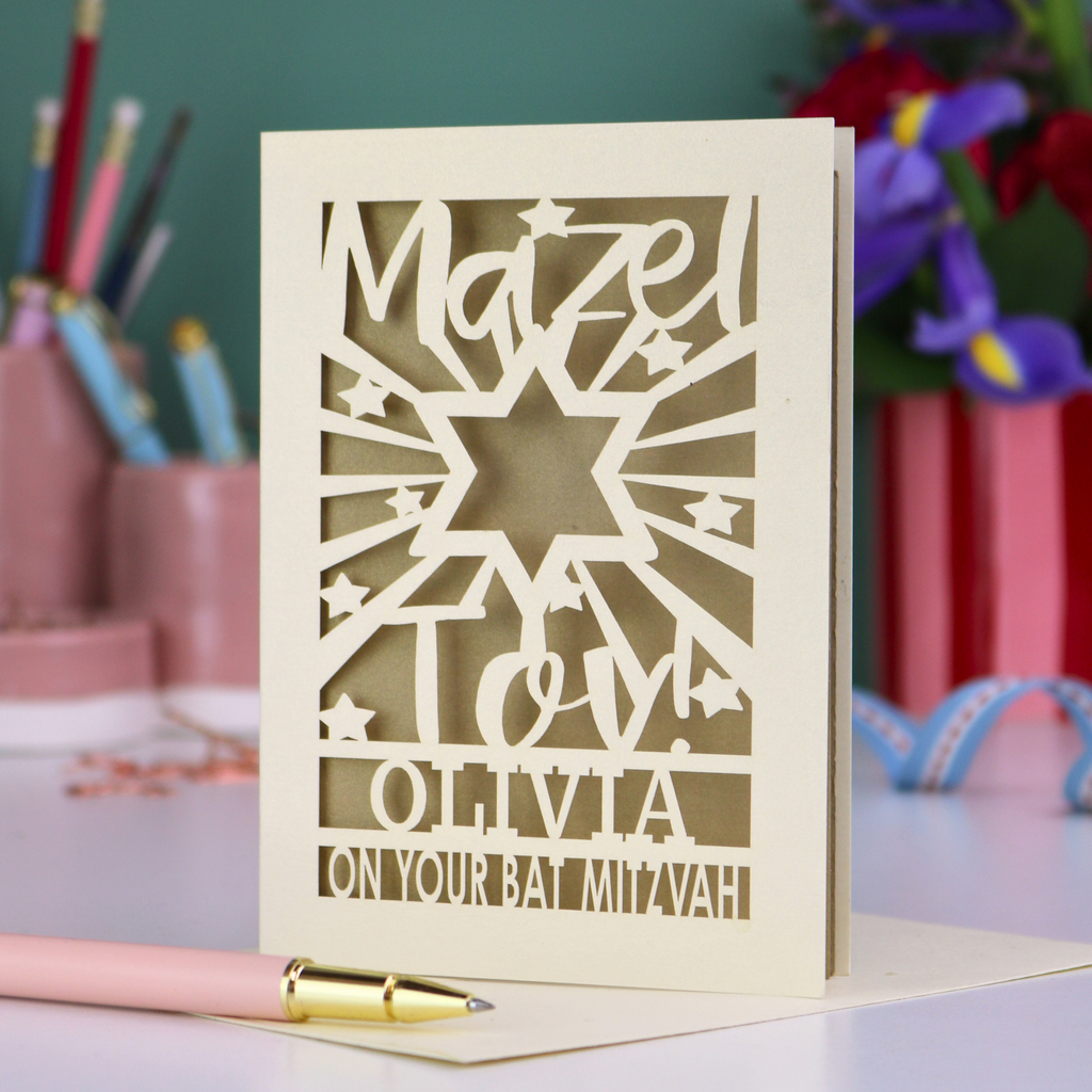 Personalized Bat Mitzvah card with decorative cut-out  star design and text 'Mazel Tov Olivia on your Bat Mitzvah' on a desk with stationery items.