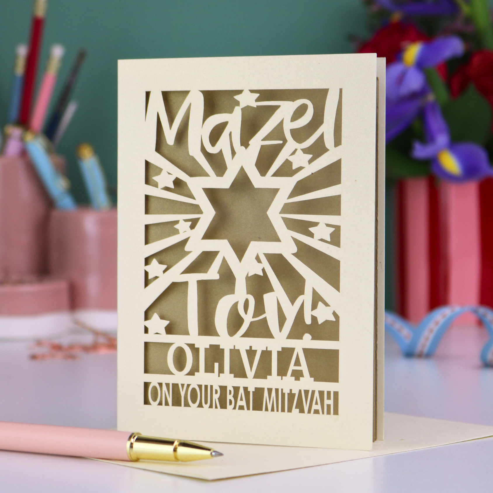 Personalized Bat Mitzvah card with decorative cut-out  star design and text 'Mazel Tov Olivia on your Bat Mitzvah' on a desk with stationery items.
