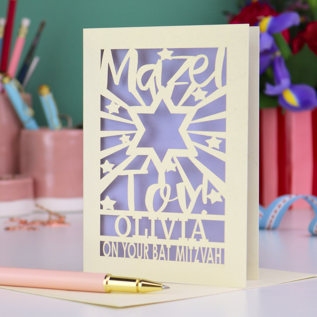 Personalized Bat Mitzvah card with decorative cut-out  star design and text 'Mazel Tov Olivia on your Bat Mitzvah' on a desk with stationery items.