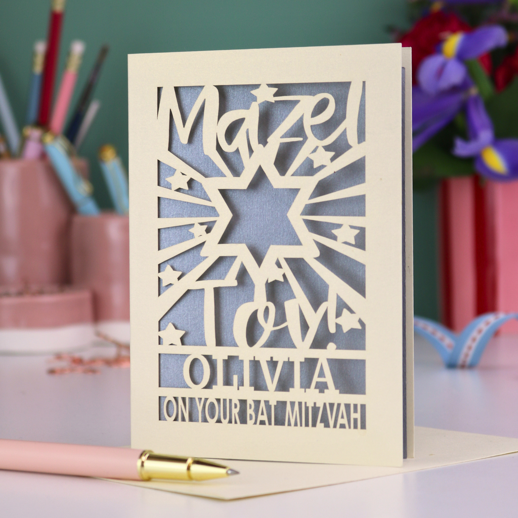 Personalized Bat Mitzvah card with decorative cut-out  star design and text 'Mazel Tov Olivia on your Bat Mitzvah' on a desk with stationery items.