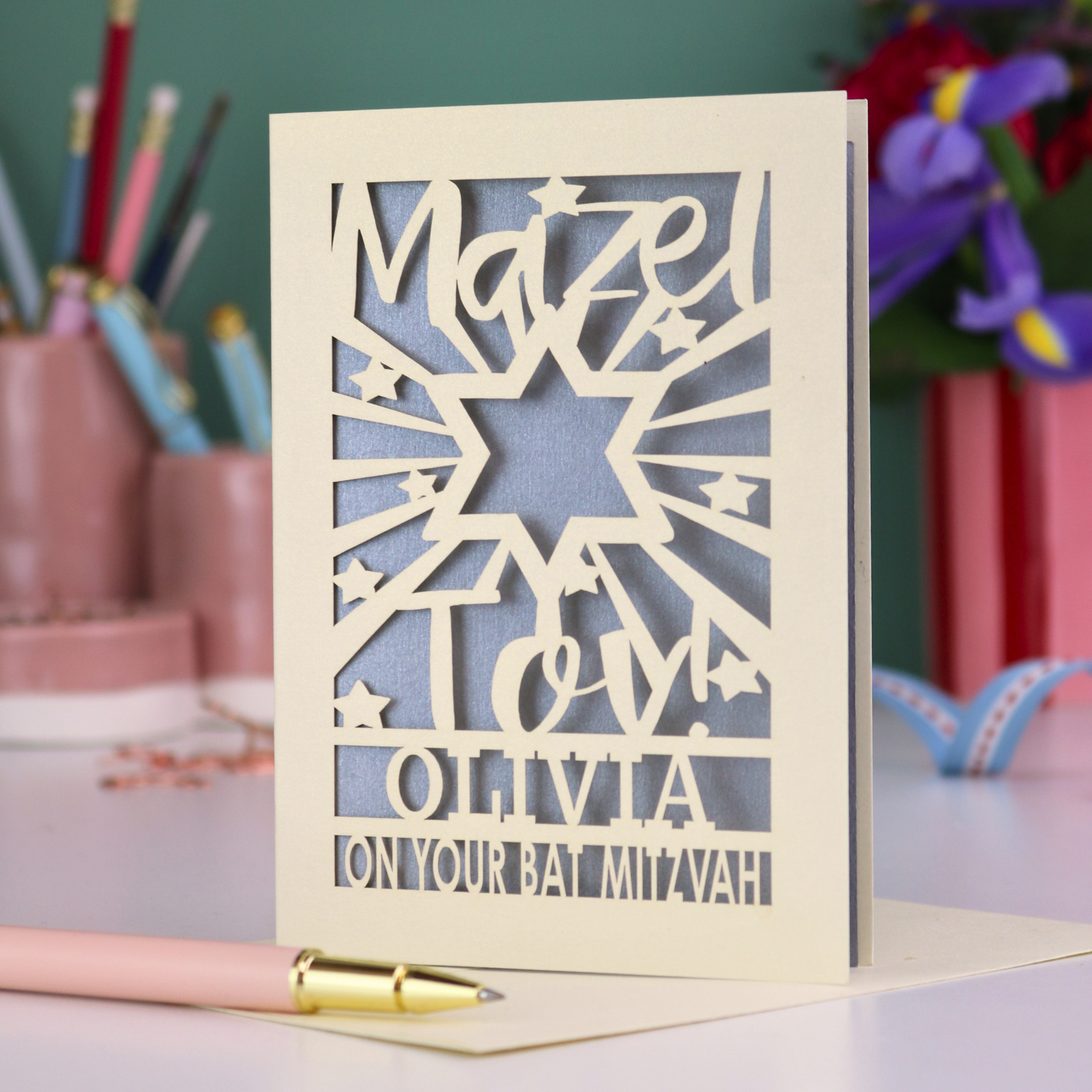 Personalized Bat Mitzvah card with decorative cut-out  star design and text 'Mazel Tov Olivia on your Bat Mitzvah' on a desk with stationery items.
