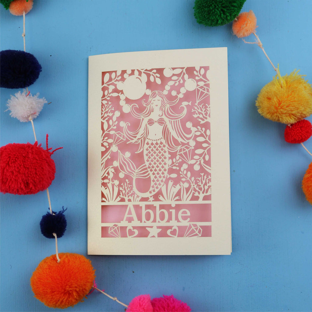 Laser cut birthday card with a mermaid and 'Abbie' on a blue background surrounded by colourful pom-poms.