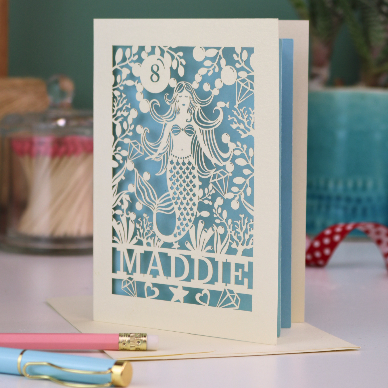 A laser cut mermaid birthday card, personalised with the age in cut out stencil font in a bubble, and the name across the bottom of the card. The design is a mermaid surrounded by bubbles, diamonds, fish and sea plants