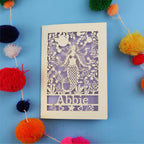 Paper cut out birthday card with a mermaid and 'Abbie' on a blue background surrounded by colourful pom-poms.
