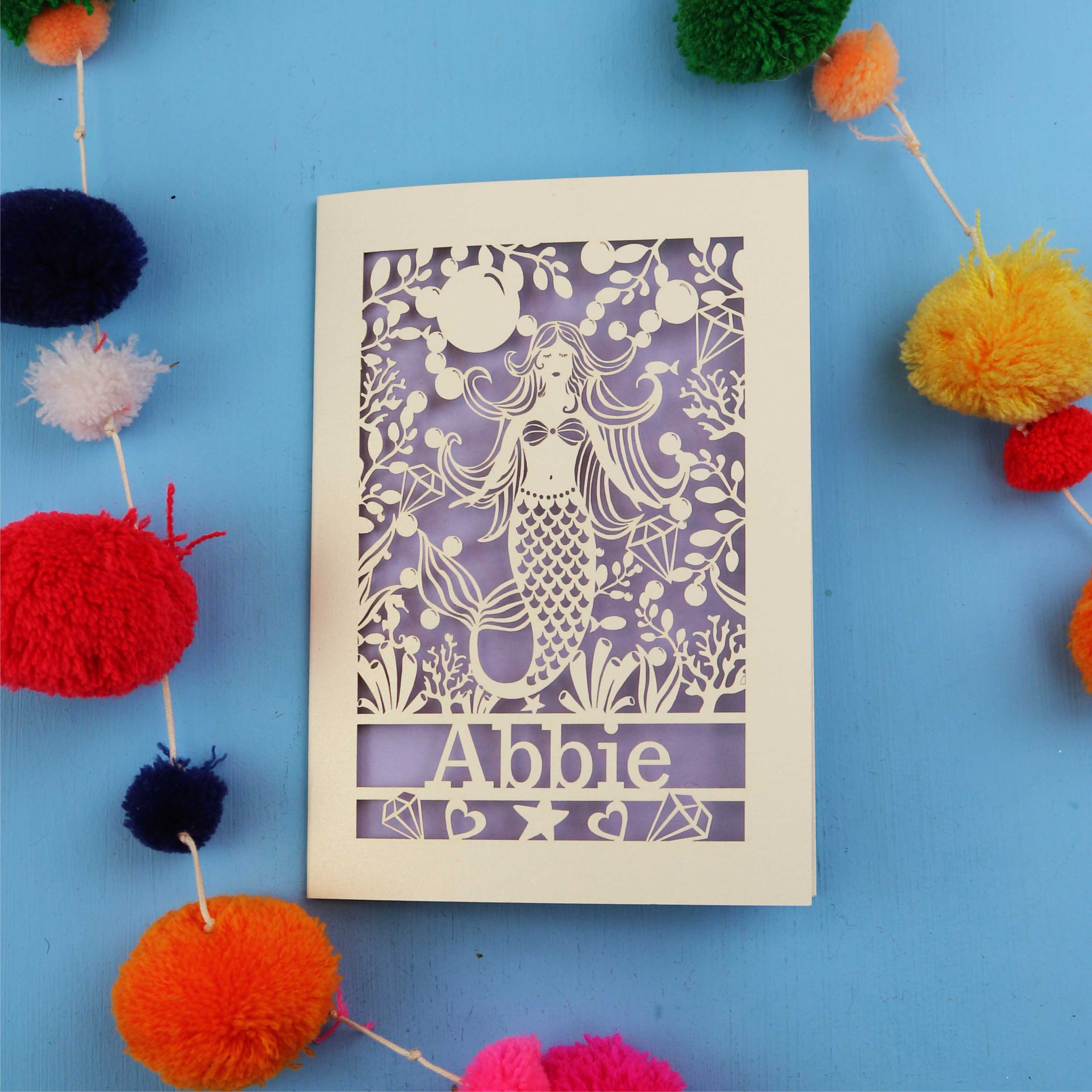 Paper cut out birthday card with a mermaid and 'Abbie' on a blue background surrounded by colourful pom-poms.