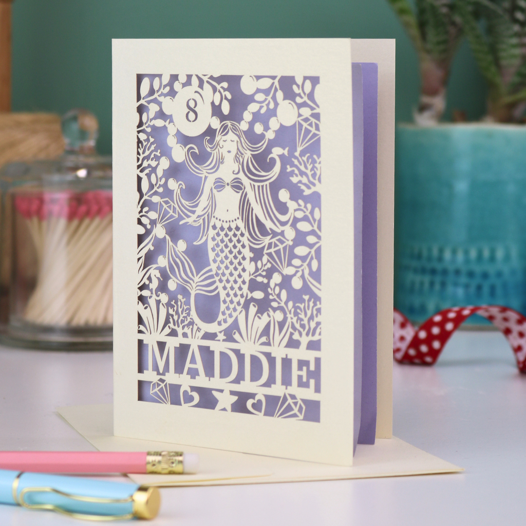 A laser cut mermaid birthday card, personalised with the age in cut out stencil font in a bubble, and the name across the bottom of the card. The design is a mermaid surrounded by bubbles, diamonds, fish and sea plants