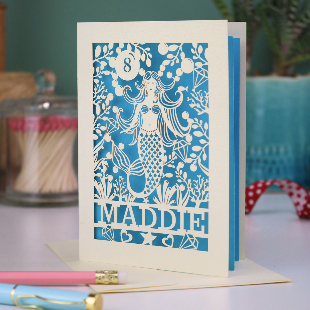 A laser cut mermaid birthday card, personalised with the age in cut out stencil font in a bubble, and the name across the bottom of the card. The design is a mermaid surrounded by bubbles, diamonds, fish and sea plants