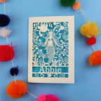 Paper cut out birthday card with a mermaid and 'Abbie' on a blue background surrounded by colourful pom-poms.