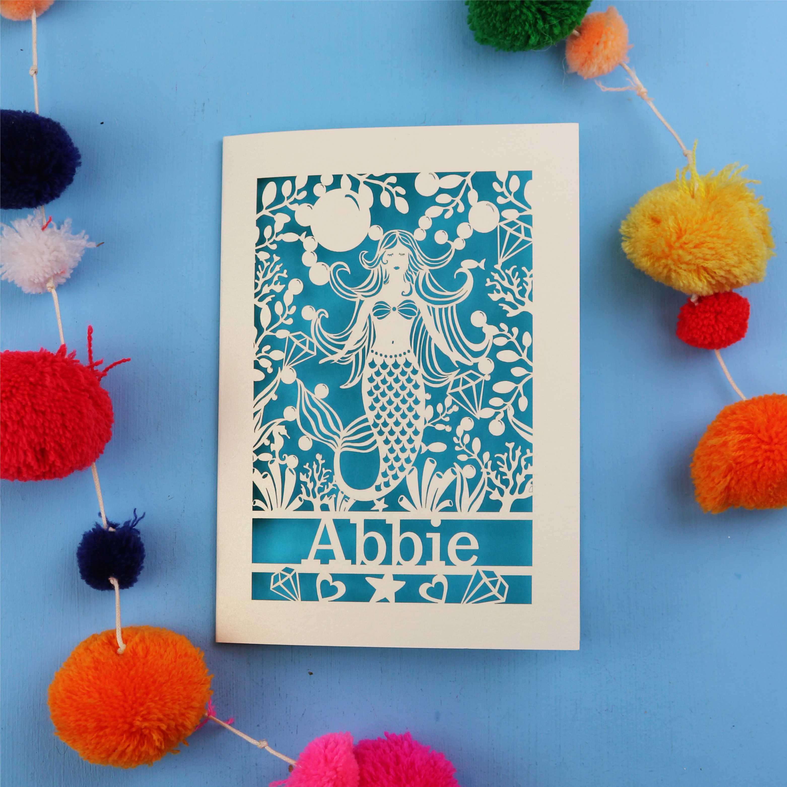 Paper cut out birthday card with a mermaid and 'Abbie' on a blue background surrounded by colourful pom-poms.