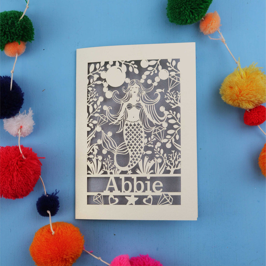 Paper cut out birthday card with a mermaid and 'Abbie' on a blue background surrounded by colourful pom-poms.