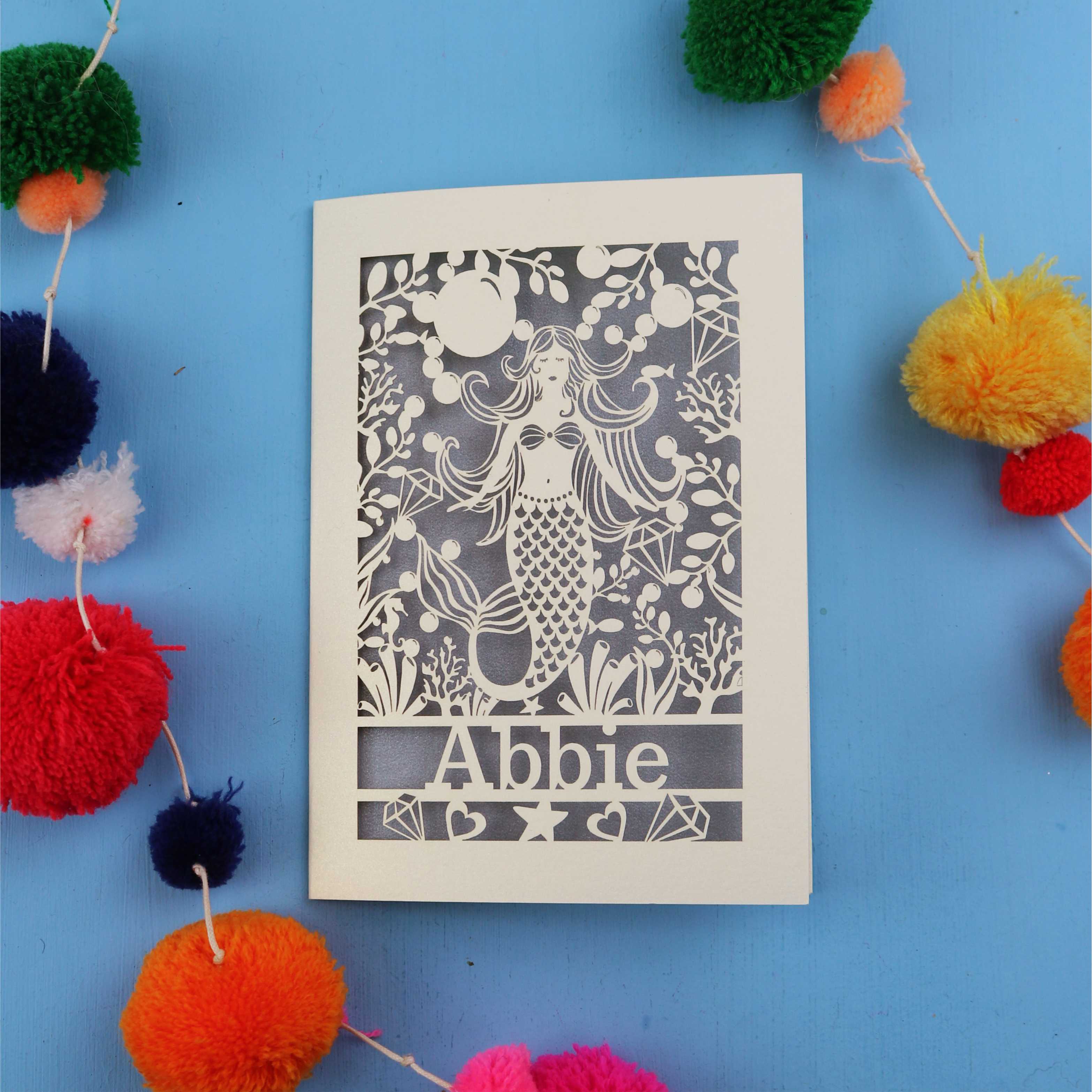 Paper cut out birthday card with a mermaid and 'Abbie' on a blue background surrounded by colourful pom-poms.