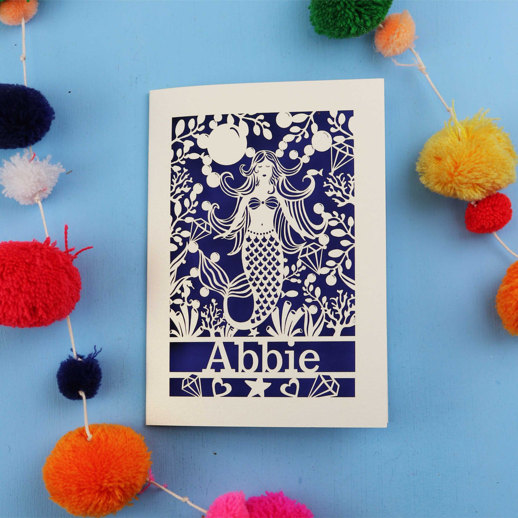 Mermaid birthday card with a mermaid and 'Abbie' on a blue background surrounded by colourful pom-poms.