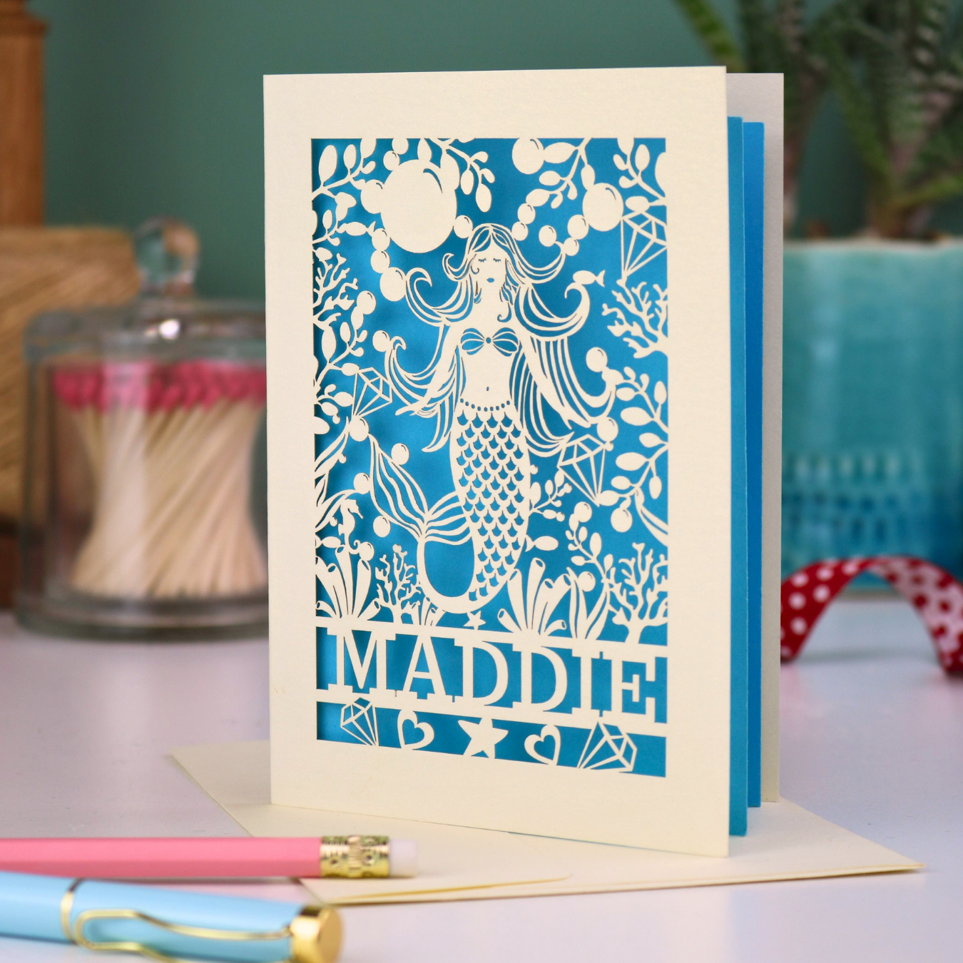 A cream laser cut birthday card with cut out mermaid design and personalised with the recipient's name. Card has a blue paper insert and stands on a white desk with stationery items.