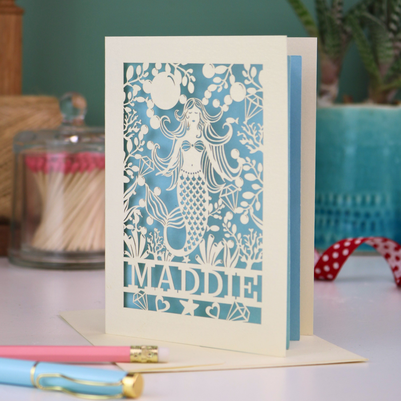 A cream laser cut birthday card with cut out mermaid design and personalised with the recipient's name. Card has a blue paper insert and stands on a white desk with stationery items.
