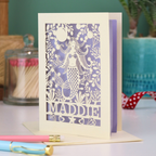 A cream laser cut birthday card with cut out mermaid design and personalised with the recipient's name. Card has a lilac paper insert and stands on a white desk with stationery items.