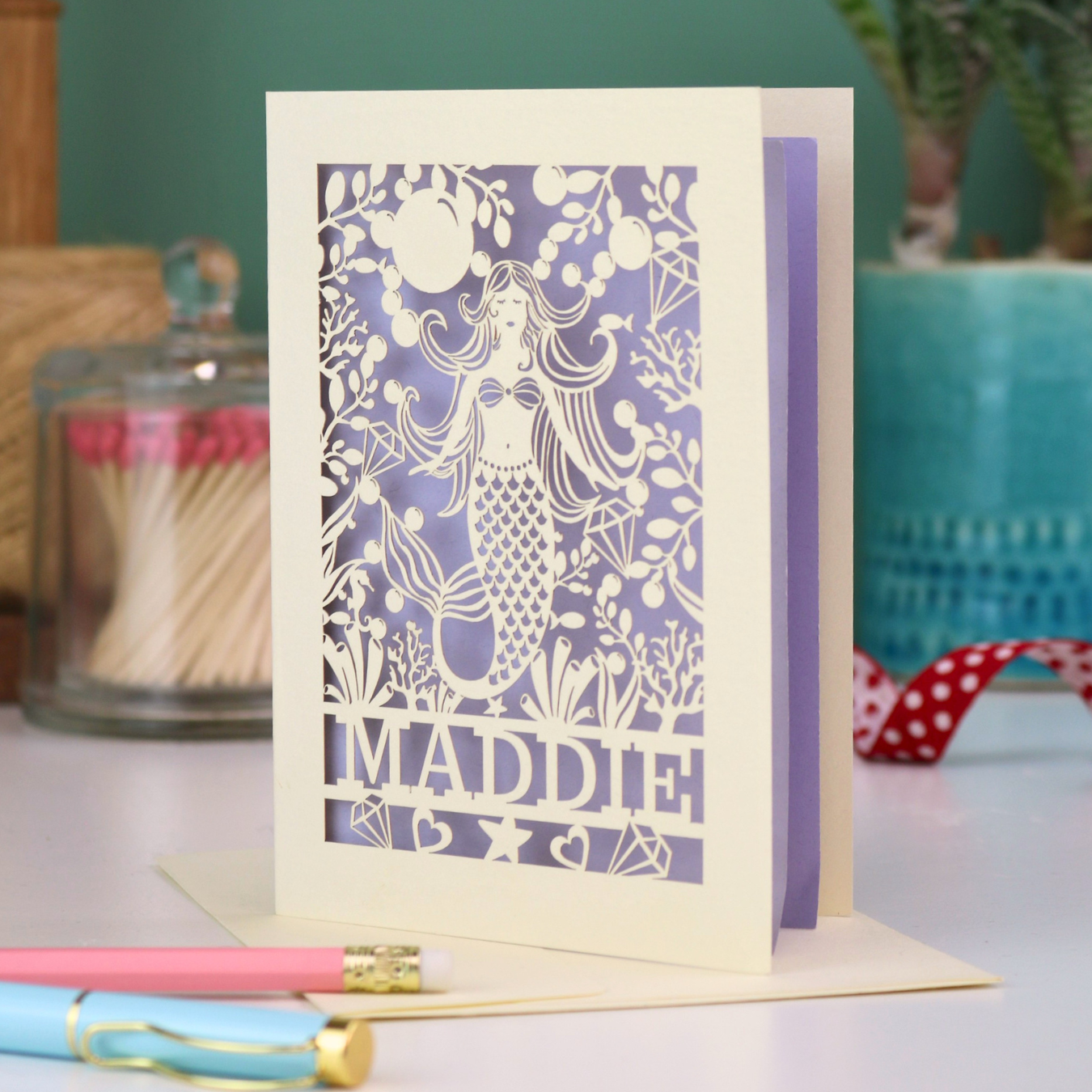 A cream laser cut birthday card with cut out mermaid design and personalised with the recipient's name. Card has a lilac paper insert and stands on a white desk with stationery items.