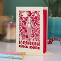A cream laser cut birthday card with cut out mermaid design and personalised with the recipient's name. Card has a shocking pink paper insert and stands on a white desk with stationery items.