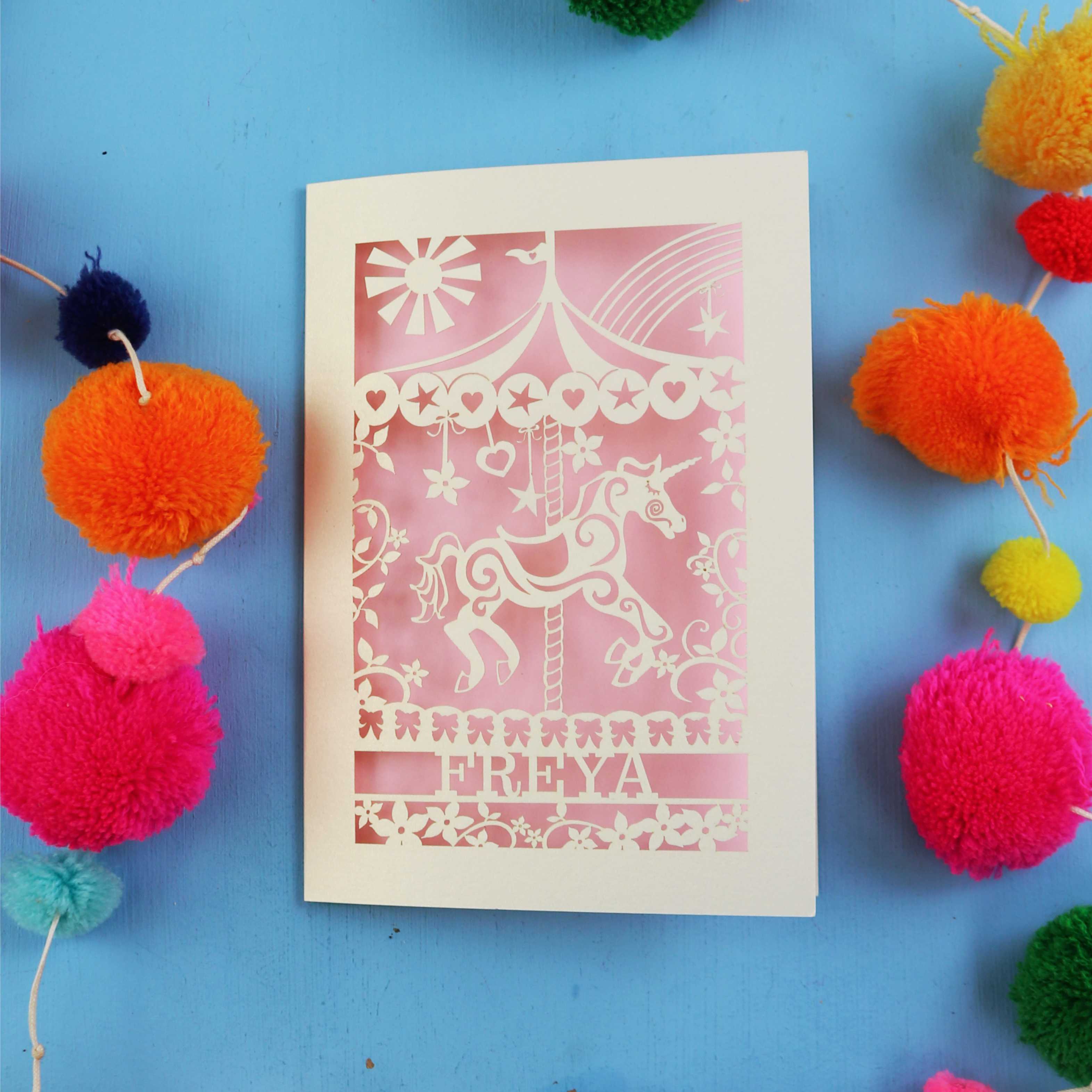 Personalised laser cut birthday Card with carousel design and 'FREYA' text on a blue background with colourful pom-poms.