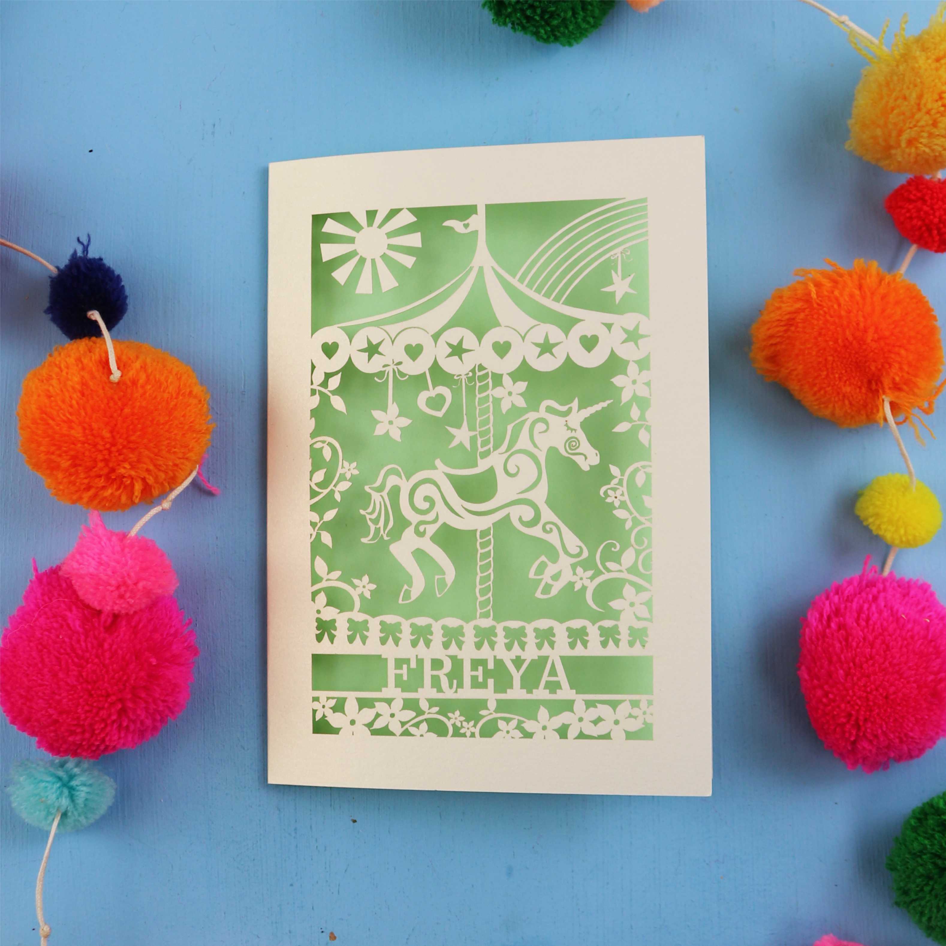 Personalised paper cut birthday Card with carousel design and 'FREYA' text on a blue background with colourful pom-poms.
