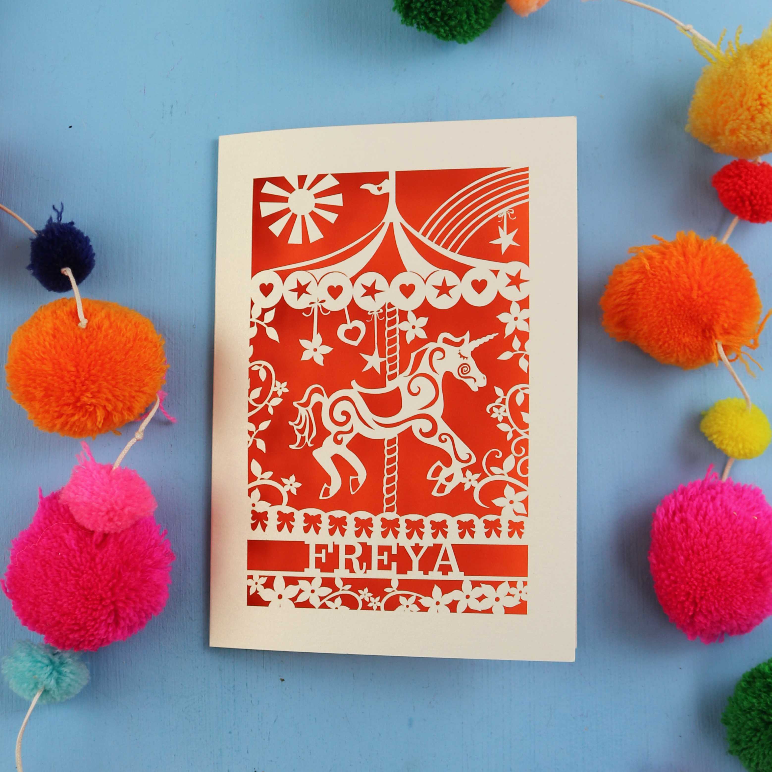 Cut out Personalised birthday Card with carousel design and 'FREYA' text on a blue background with colourful pom-poms.