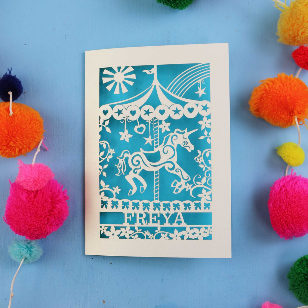 Personalised birthday Card with carousel design and 'FREYA' text on a blue background with colourful pom-poms.