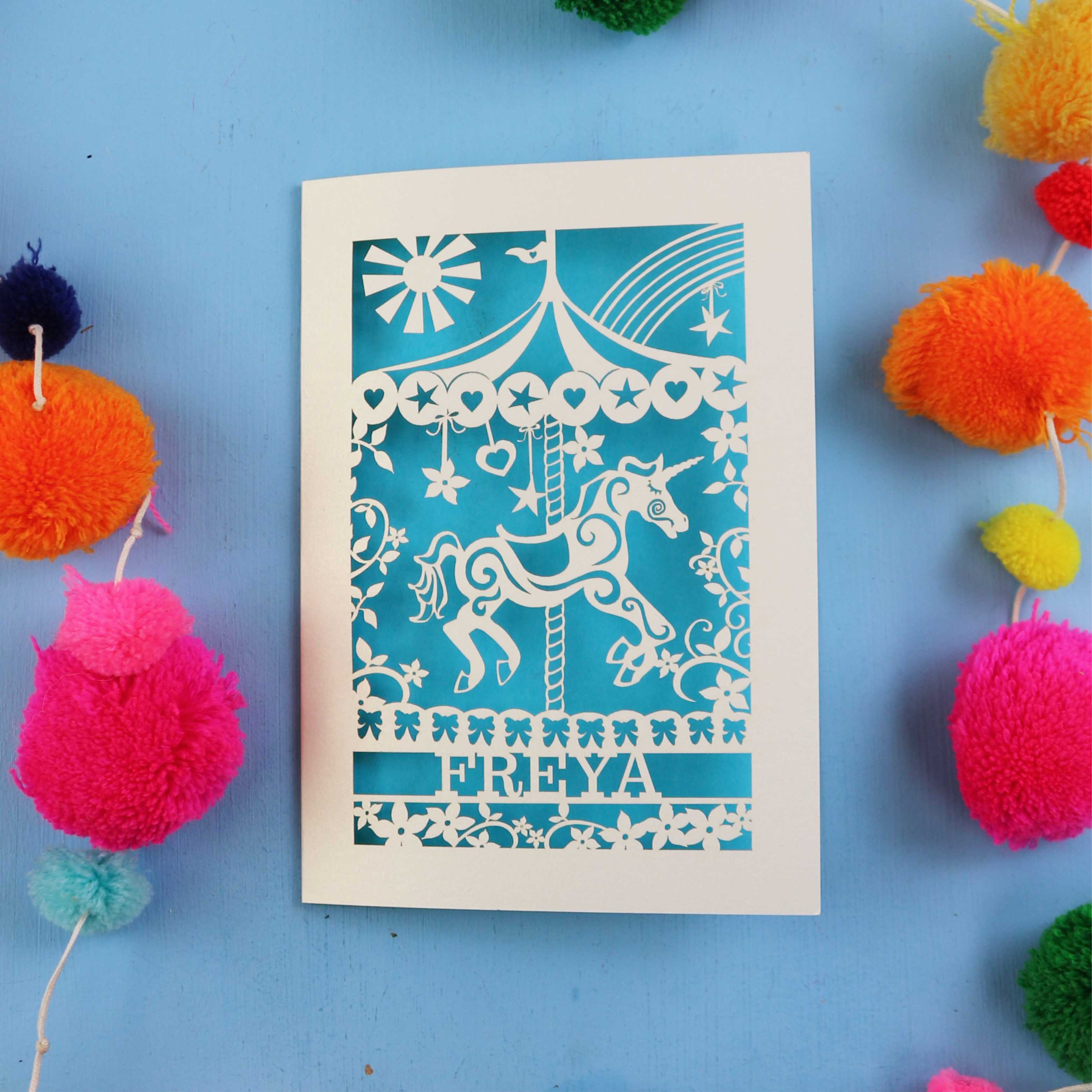 Personalised birthday Card with carousel design and 'FREYA' text on a blue background with colourful pom-poms.