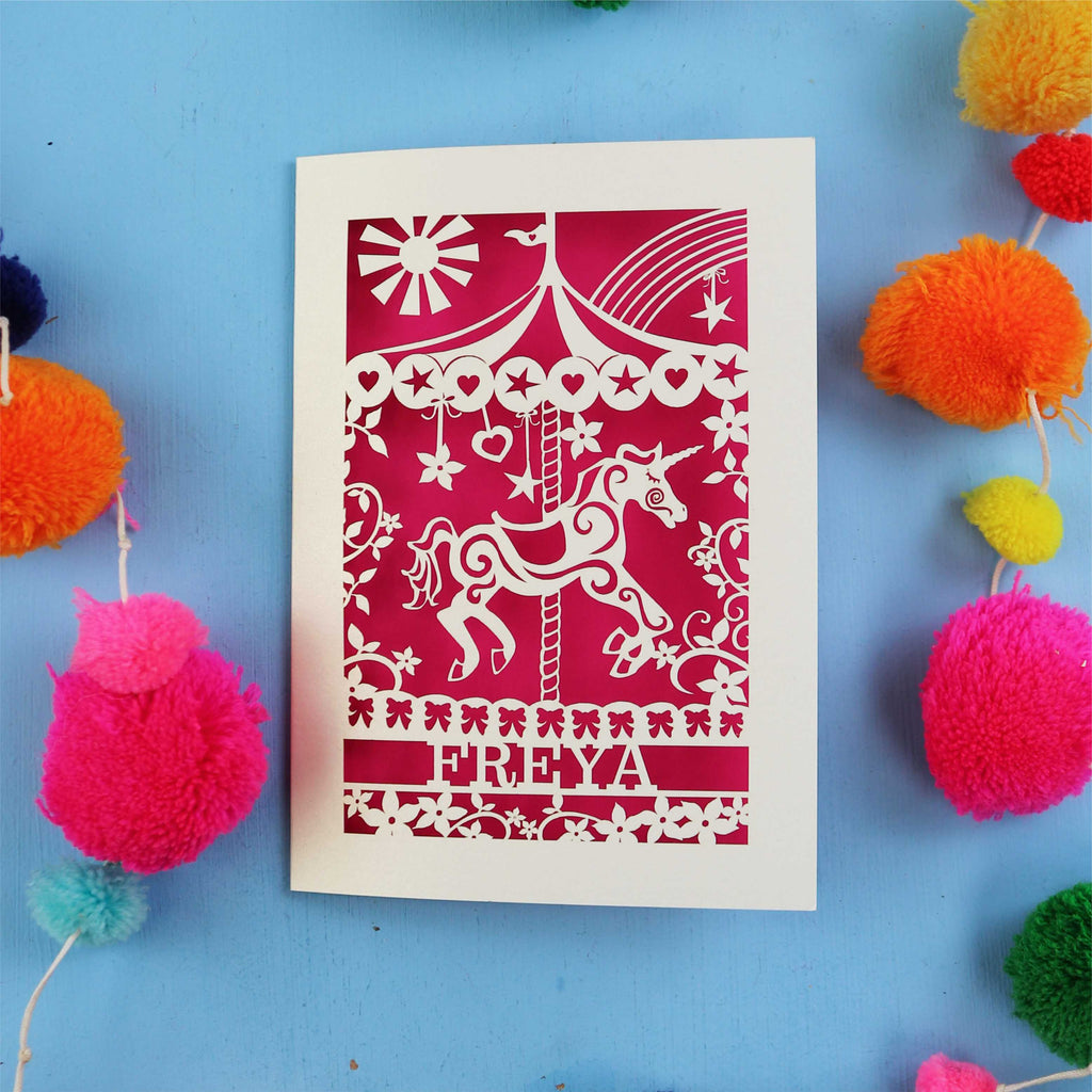 Personalised birthday Card with carousel design and 'FREYA' text on a blue background with colourful pom-poms.