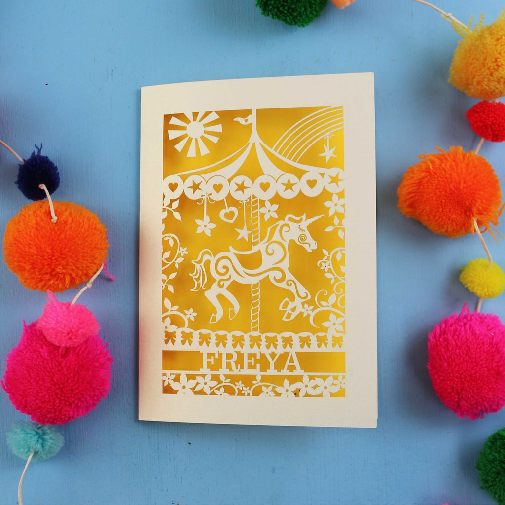 Personalised birthday Card with carousel design and 'FREYA' text on a blue background with colourful pom-poms.