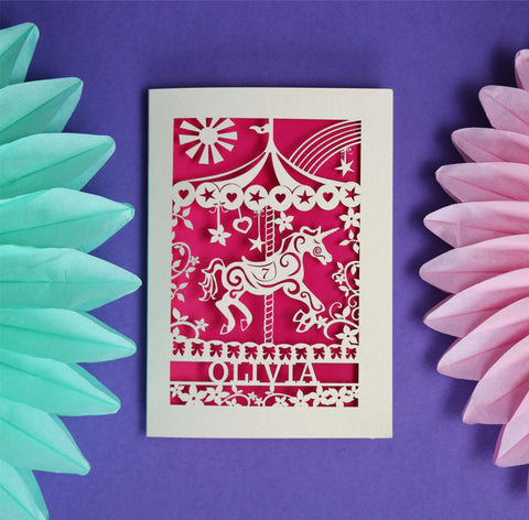 A laser cut birthday card personalised with a name and an age. Card features a carousel horse surrounded by flowers, a rainbow and the sun.  - 