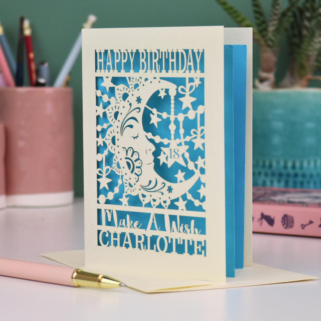 A personalised Birthday card with laser cut crescent moon design and text, 'Happy Birthday, Make a Wish Charlotte', on a desk with stationery items.