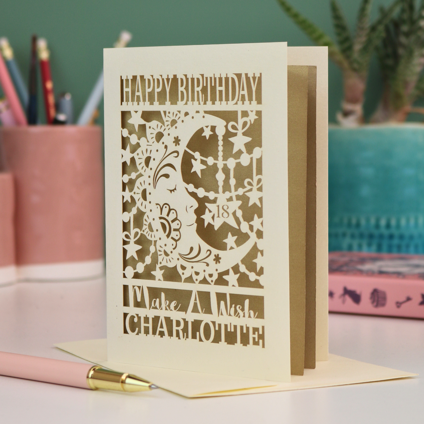 A personalised Birthday card with laser cut crescent moon design and text, 'Happy Birthday, Make a Wish Charlotte', on a desk with stationery items.
