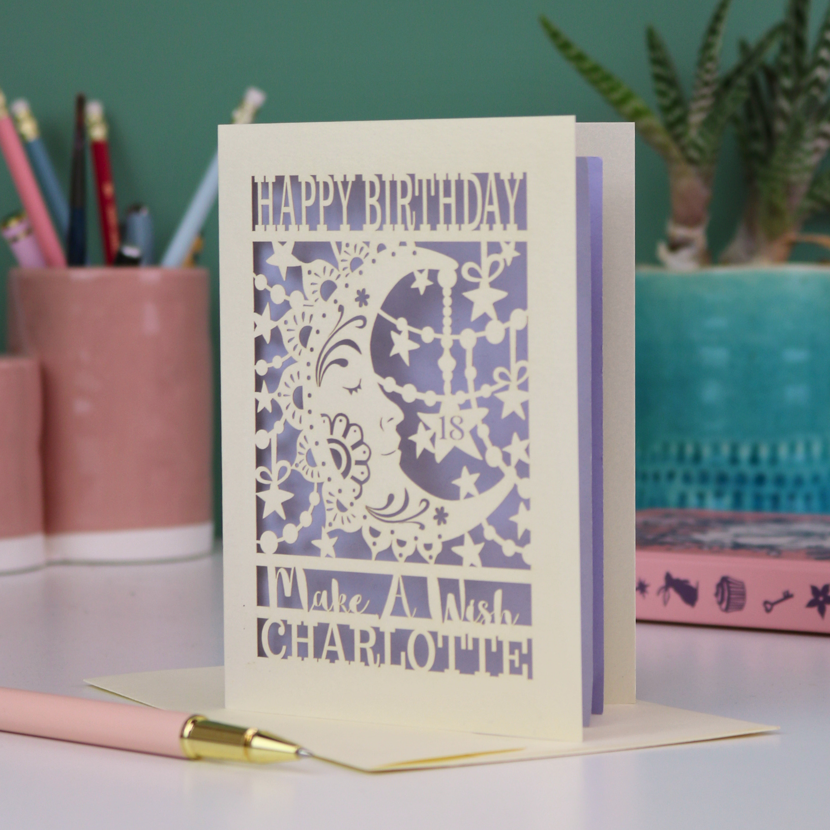 A personalised Birthday card with laser cut crescent moon design and text, 'Happy Birthday, Make a Wish Charlotte', on a desk with stationery items.