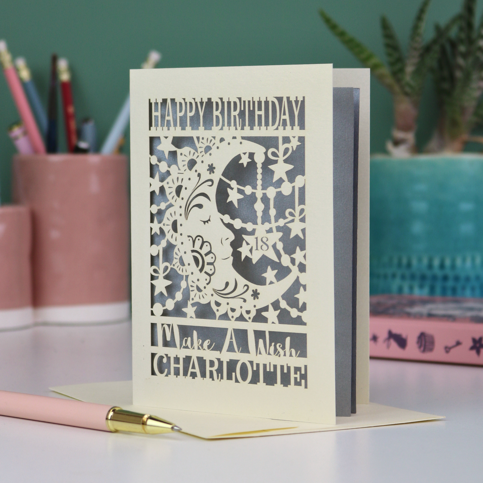 A personalised Birthday card with laser cut crescent moon design and text, 'Happy Birthday, Make a Wish Charlotte', on a desk with stationery items.