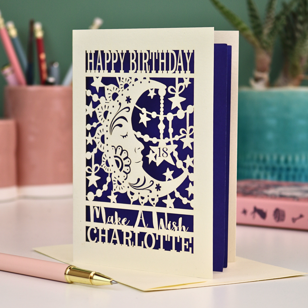 A personalised Birthday card with laser cut crescent moon design and text, 'Happy Birthday, Make a Wish Charlotte', on a desk with stationery items.
