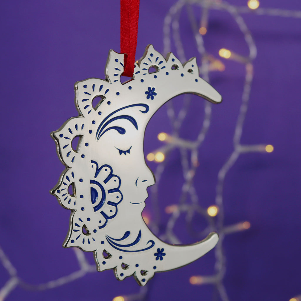 A silver-coloured crescent moon Christmas decoration with intricate patterns, hanging from a red ribbon against a purple background with blurred lights.