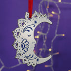 A silver-coloured crescent moon Christmas decoration with intricate patterns, hanging from a red ribbon against a purple background with blurred lights.