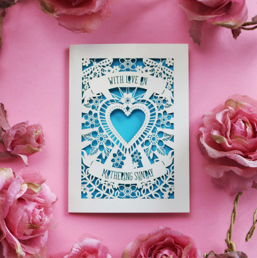 A cream laser cut Mothering Sunday card that says "With love on Mothering Sunday". Card has a heart and flower details and a peacock blue paper insert