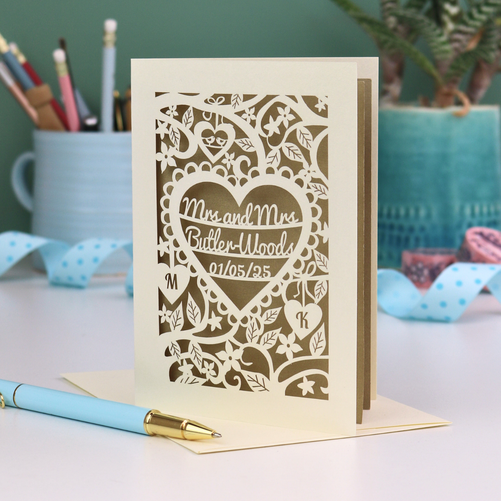 Laser-cut Mrs and Mrs anniversary card with personalized text, 'Mrs and Mrs Butler-Woods, 01/05/25', surrounded by leaves and flowers,  on a desk with stationery items.