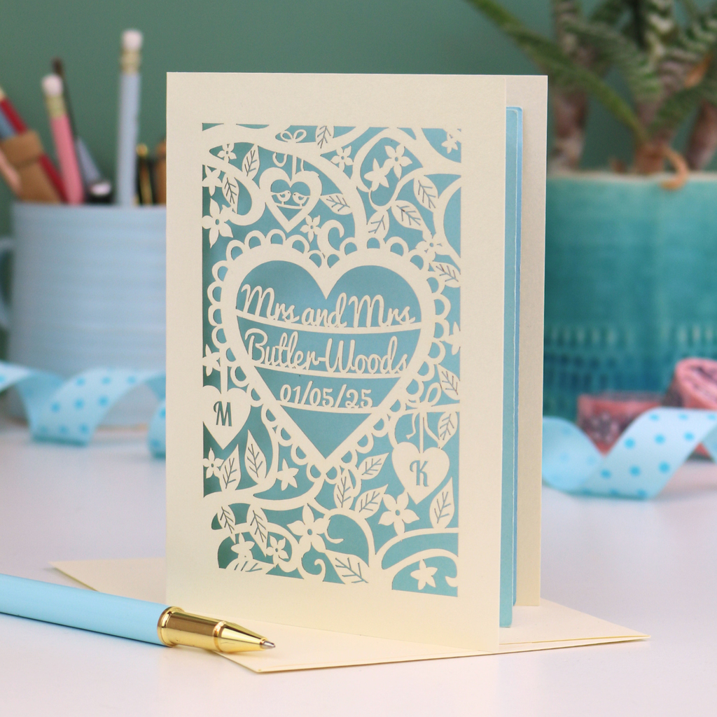 Laser-cut gay anniversary card with personalized text, 'Mrs and Mrs Butler-Woods, 01/05/25', surrounded by leaves and flowers,  on a desk with stationery items.