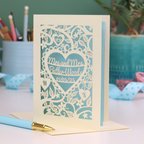Laser-cut gay anniversary card with personalized text, 'Mrs and Mrs Butler-Woods, 01/05/25', surrounded by leaves and flowers,  on a desk with stationery items.
