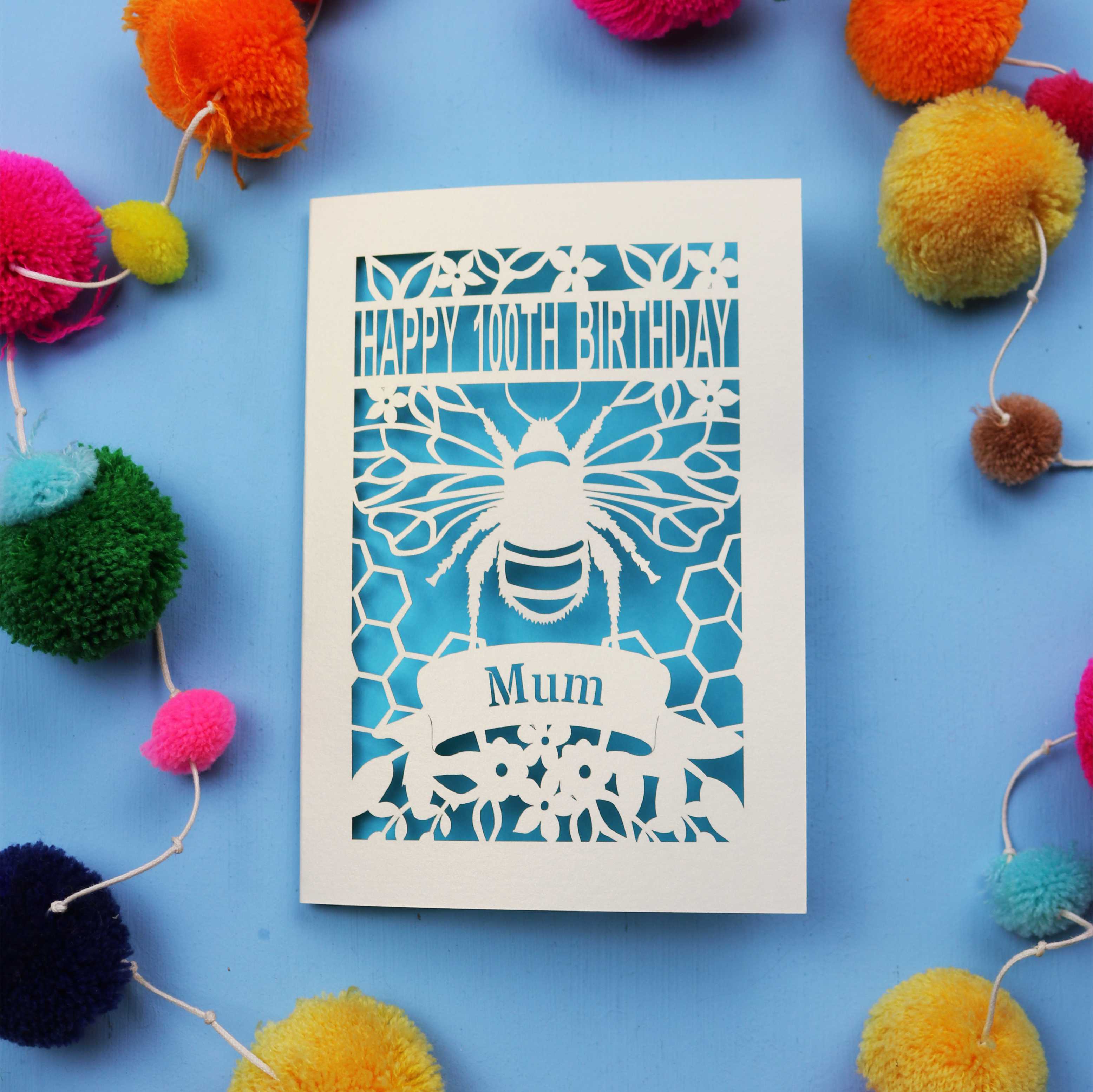 Paper cut 100th birthday card for 'Mum' with bee design on a blue background with colourful pom-poms.