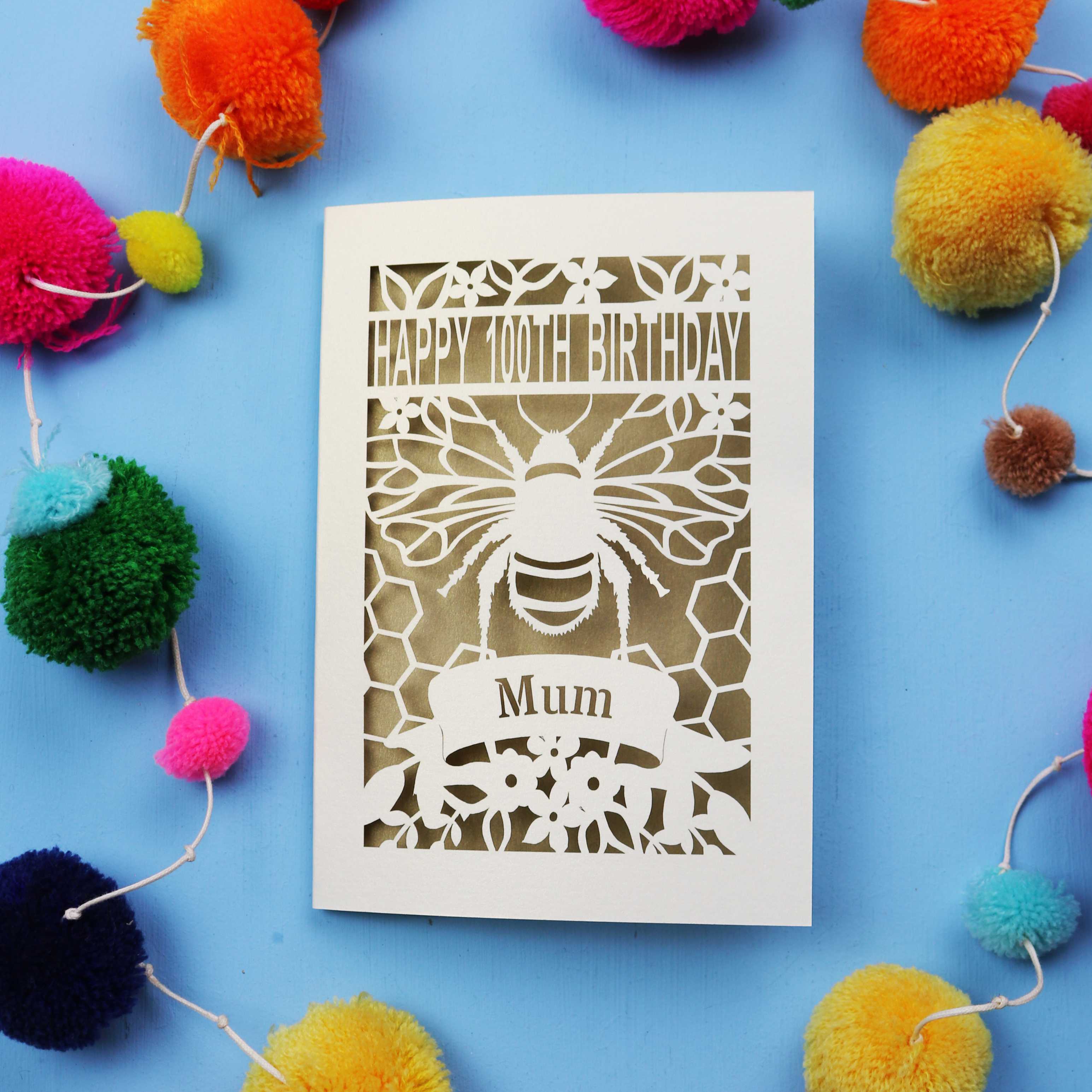 Laser-cut 100th birthday card for 'Mum' with bee design on a blue background with colourful pom-poms.