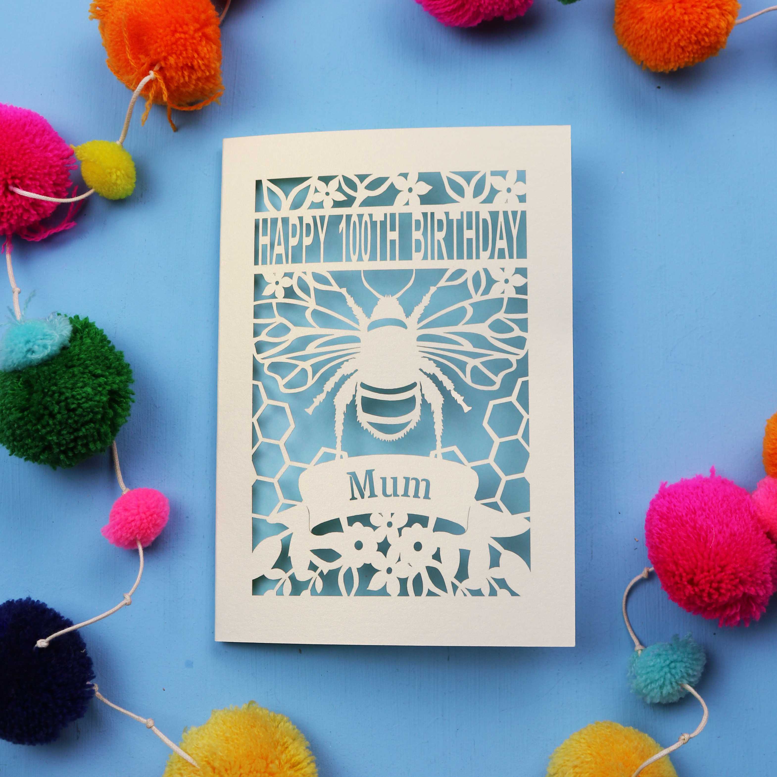 Laser-cut 100th birthday card for 'Mum' with bee design on a blue background with colourful pom-poms.