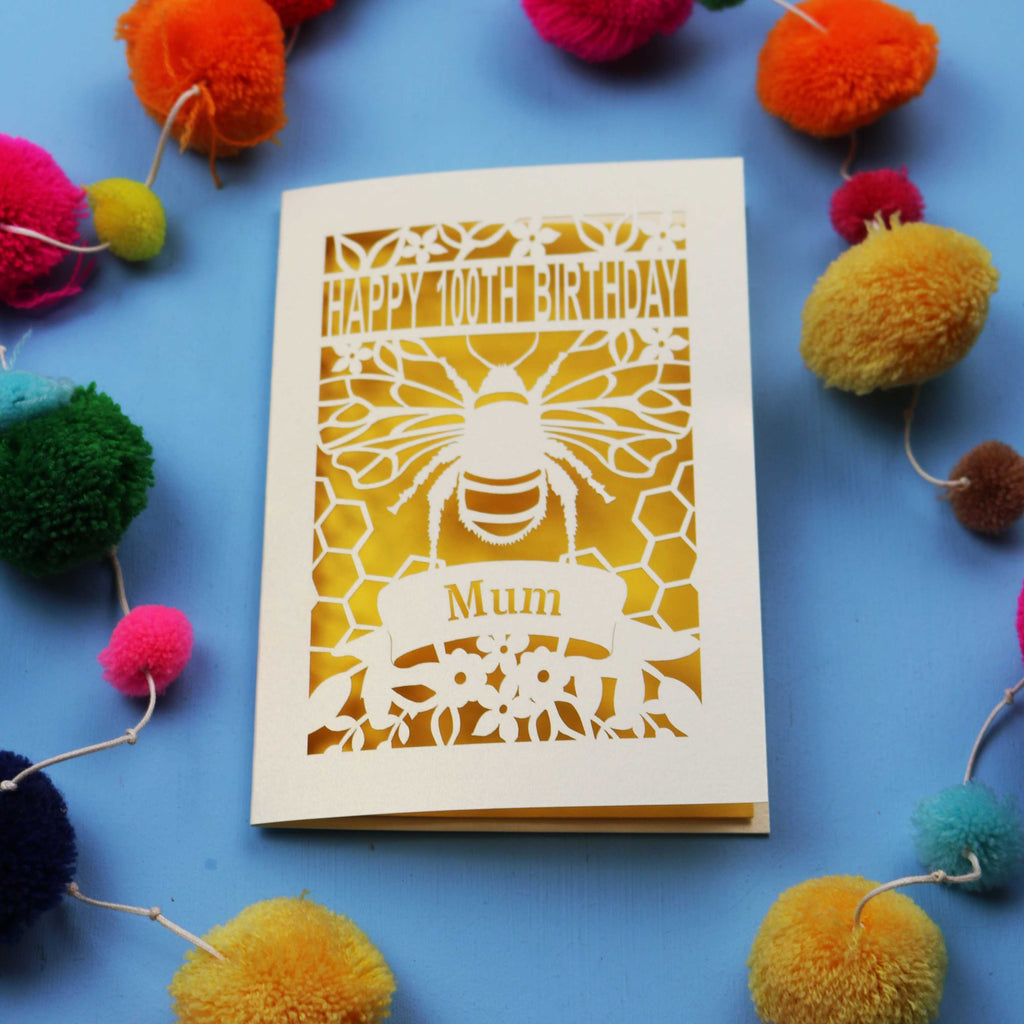 Laser-cut 100th birthday card for 'Mum' with bee design on a blue background with colourful pom-poms.