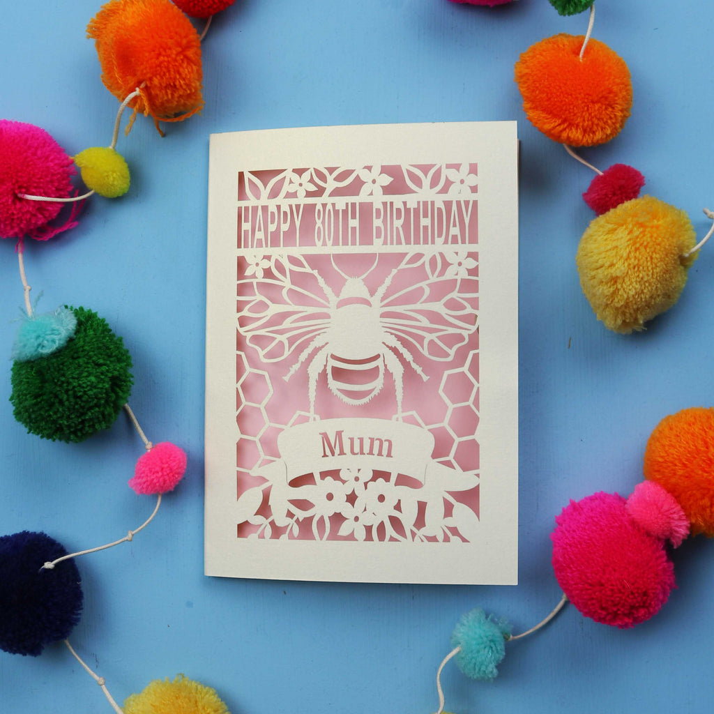 Laser-cut birthday card with bee design and 'Happy 80th Birthday Mum' text on a blue background with colourful pom-poms.